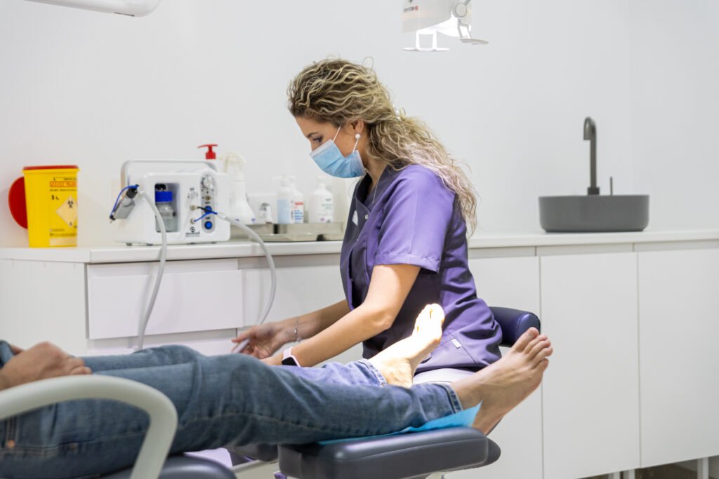 Podóloga con uniforme azul y mascarilla atendiendo los pies de un paciente recostado en la silla de tratamiento dentro del consultorio.