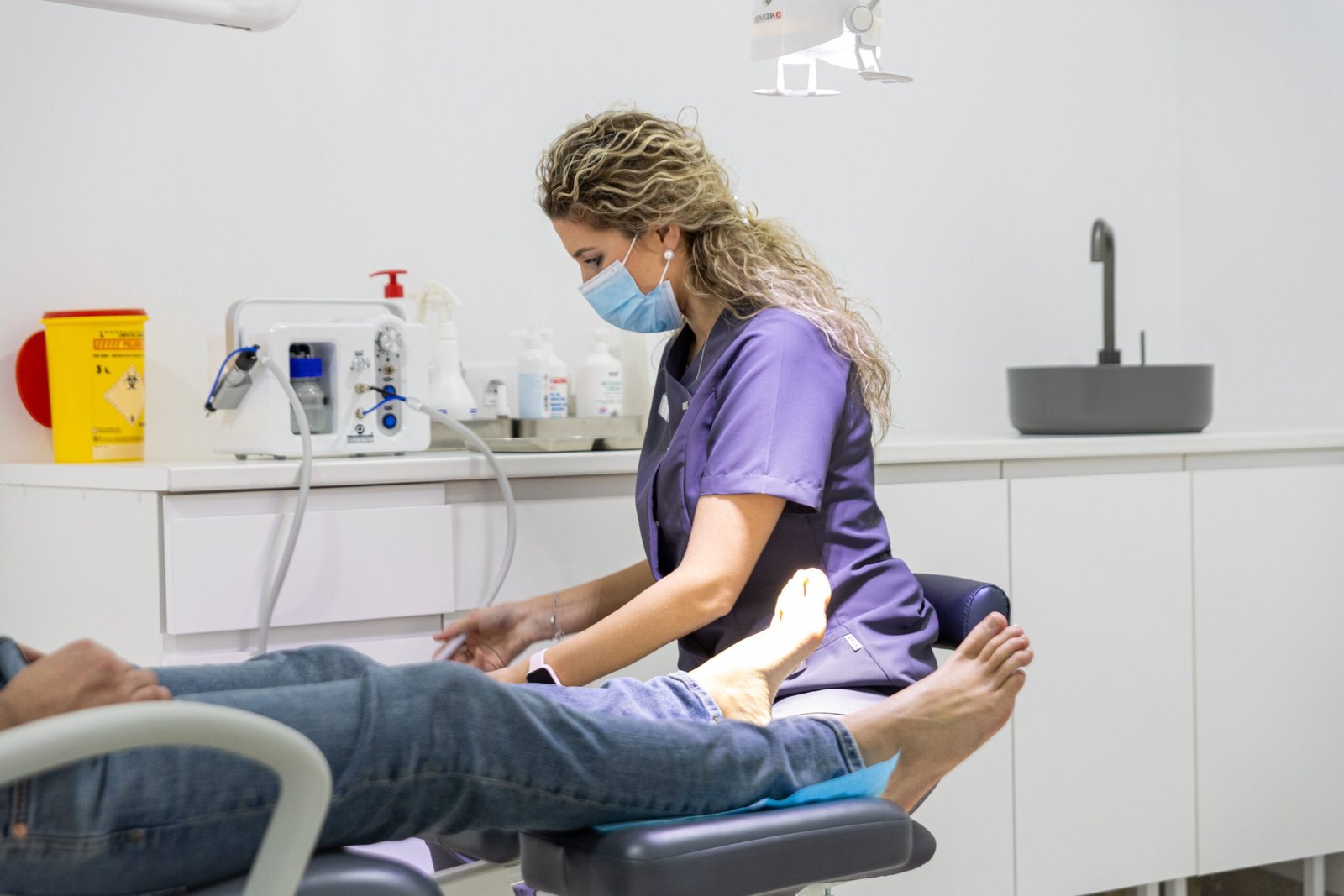 Podóloga con uniforme azul y mascarilla atendiendo los pies de un paciente recostado en la silla de tratamiento dentro del consultorio.