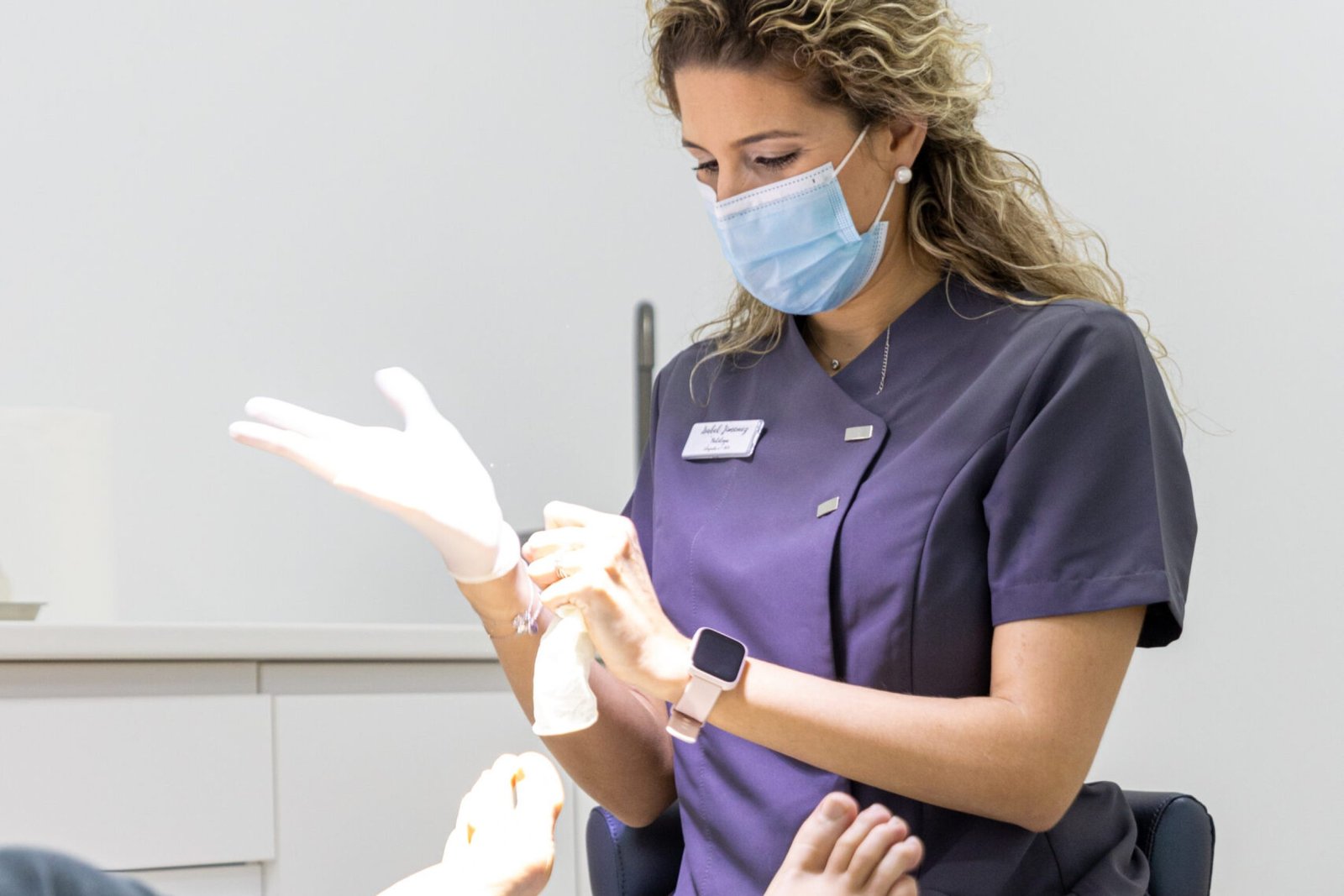 Podóloga con uniforme azul y mascarilla colocándose guantes de látex mientras un paciente está recostado en una silla de tratamiento.